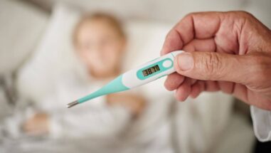 Zum Artikel: Fieberthermoter in der Hand und im Hintergrund ein Kind